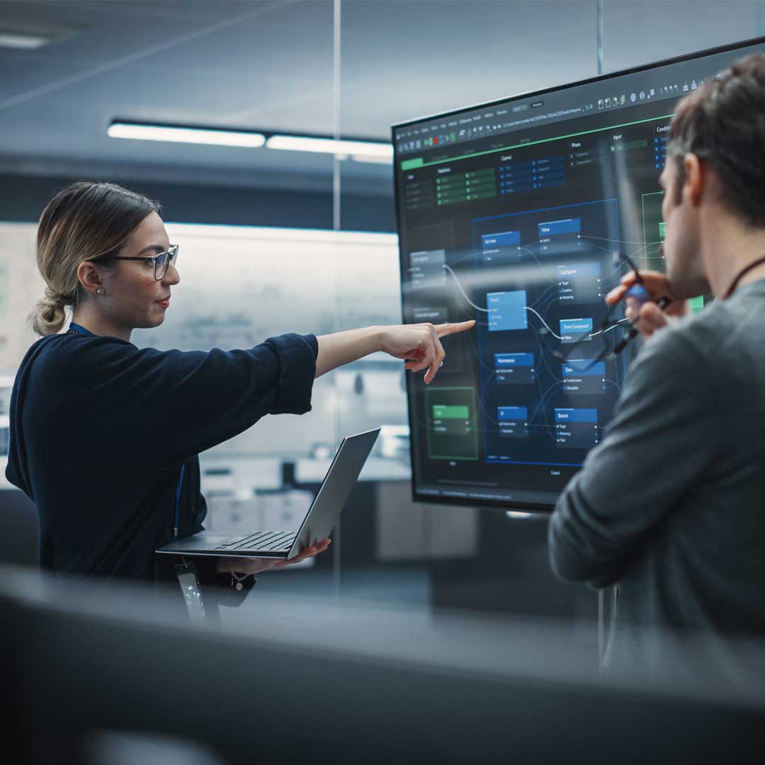 Eine Mitarbeiterin eines Tech-Unternehmens zeigt ihrem Kollegen auf einem großen Bildschirm Automatisierungen.