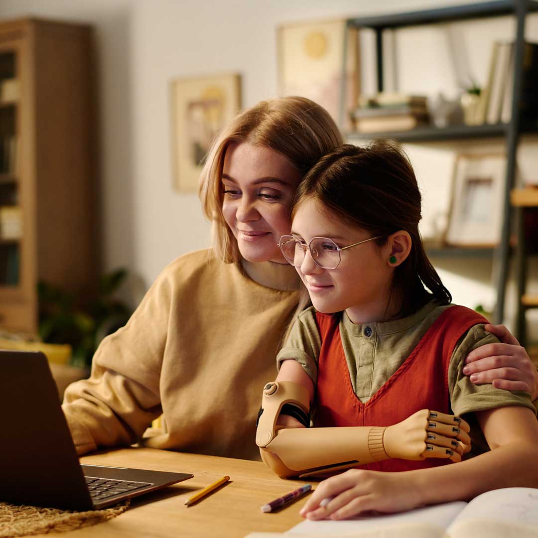 Eine Frau und ein junges Mädchen mit einer Armprothese sitzen an einem Tisch mit einem Laptop.