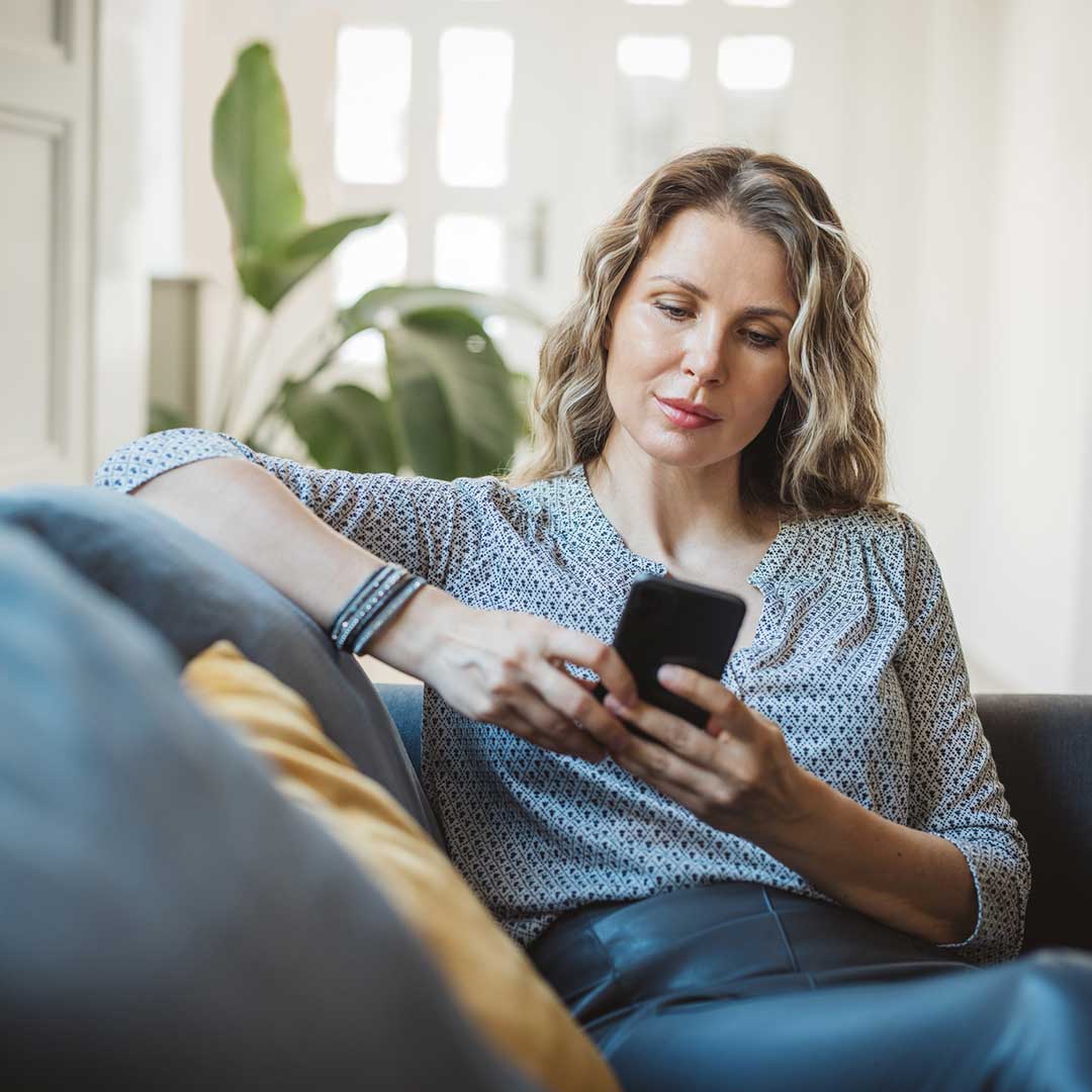 Ein blonde Frau sitzt auf dem Sofa und tippt auf ihrem Smartphone