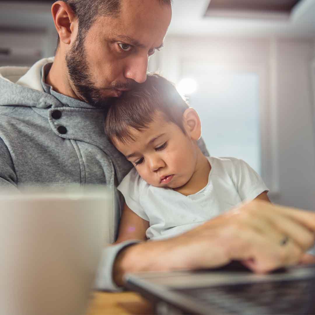 Mann sitzt am Laptop und hält ein kleines Kind im Arm.