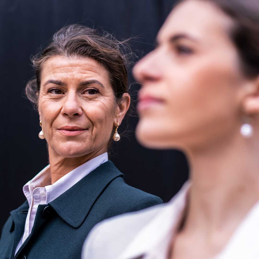 Eine schick gekleidete ältere Unternehmerin steht im Hintergrund und blickt auf eine junge Mitarbeiterin im Vordergrund.