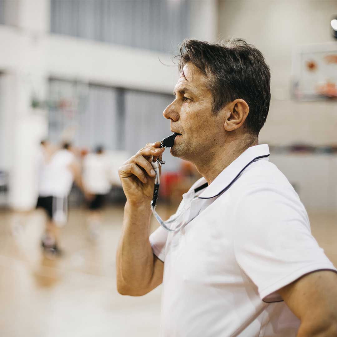 Männlicher Trainer im weißen Polohemd mit Pfeife im Mund steht auf dem Basketballfeld.