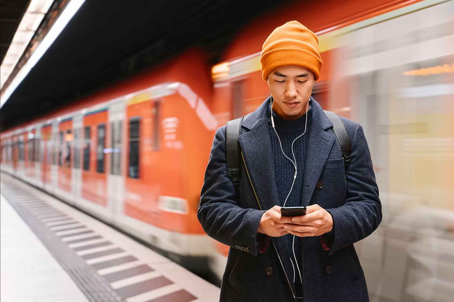 Mann mit Mütze steht am Bahnhof und blickt in sein Smartphone