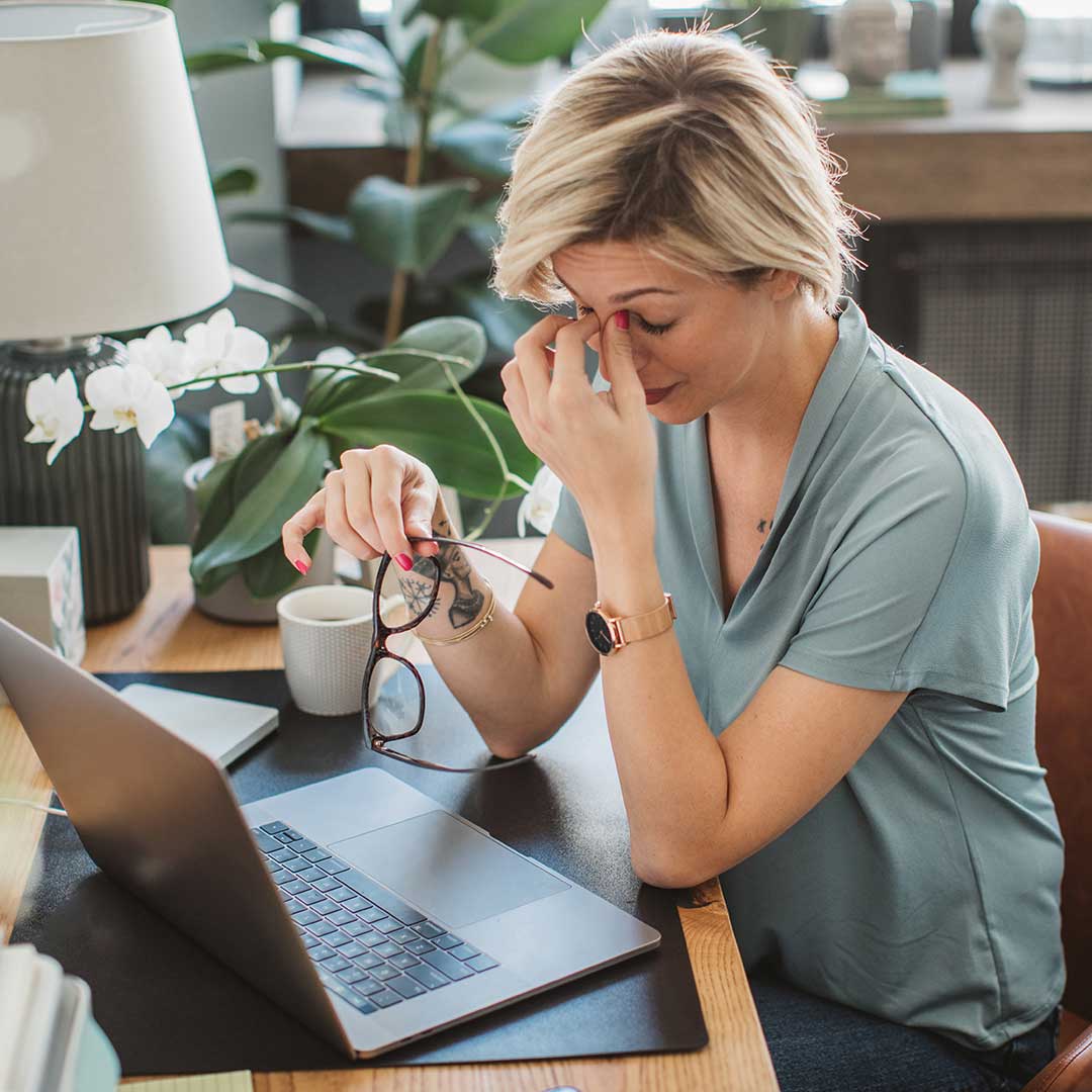 Junge Frau sitzt am Schreibtisch vor ihrem Laptop und reibt sich müde die Augen