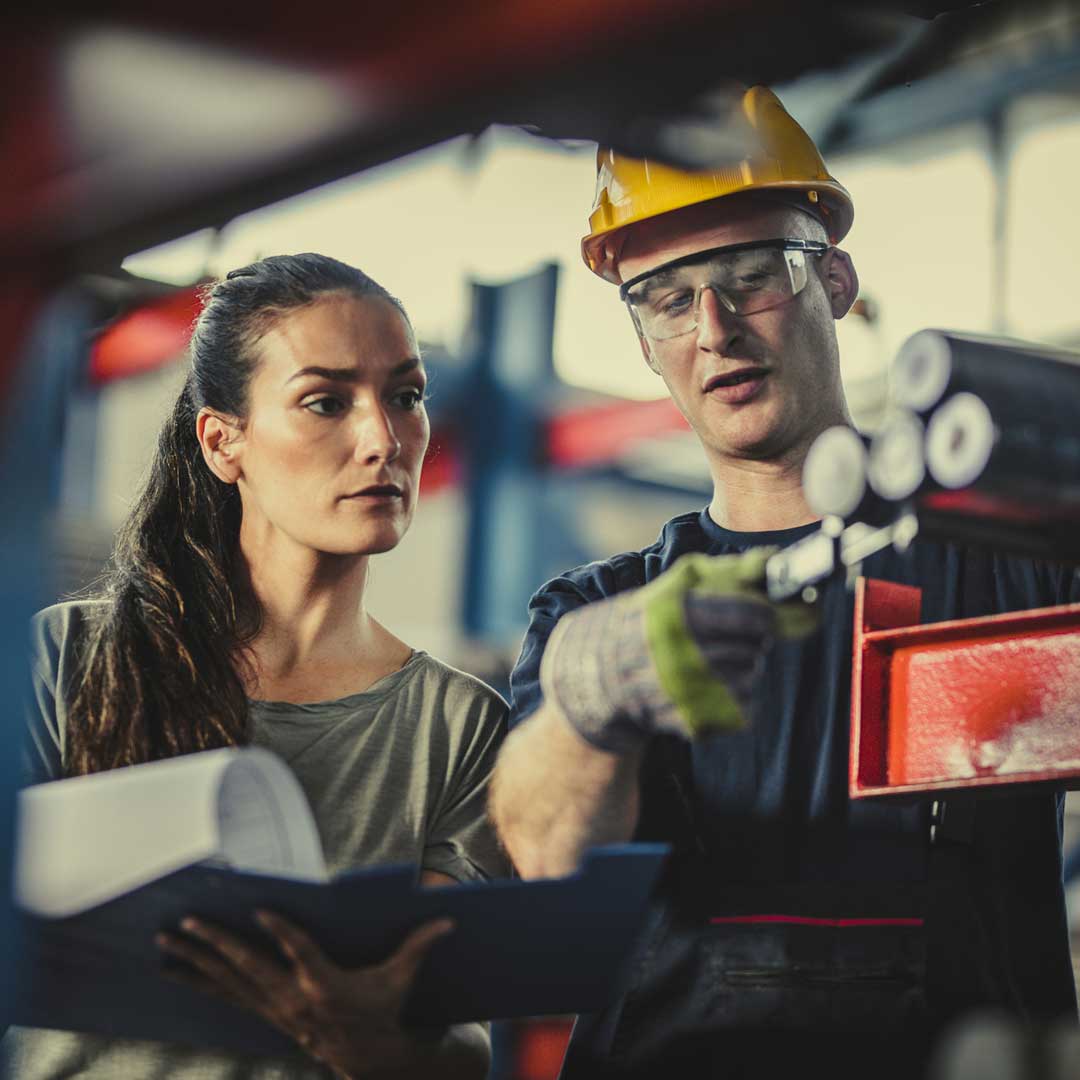 Zwei Personen in einer Fabrikumgebung prüfen metallische Zylinderstangen. Der Mann mit gelbem Schutzhelm misst eine Stange mit einem Messschieber, während die andere weibliche Person mit einem blauen Klemmbrett daneben steht und den Vorgang beobachtet