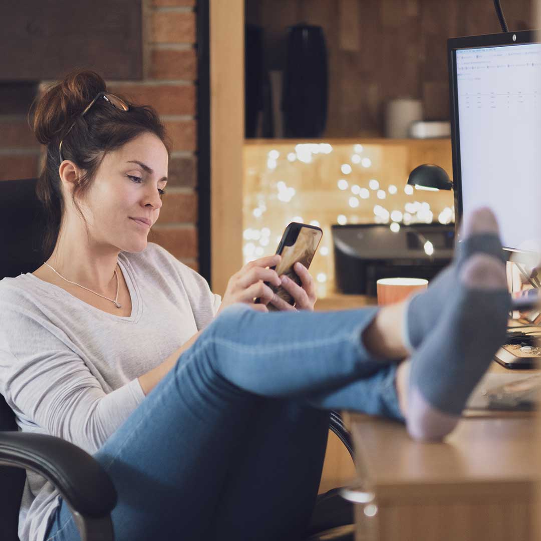 Frau sitzt entspannt am Schreibtisch mit ihren Füßen auf dem Tisch und hält ein Smartphone in den Händen